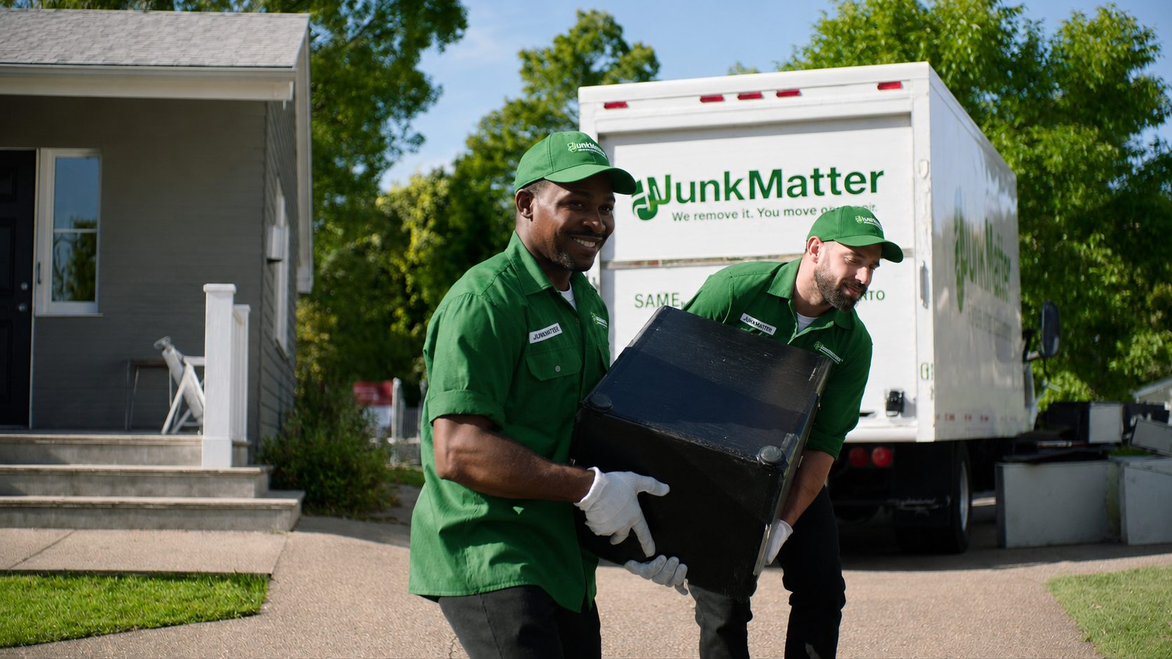 Two smiling JunkMatter crew members in branded uniforms carrying a cabinet from a house to their truck